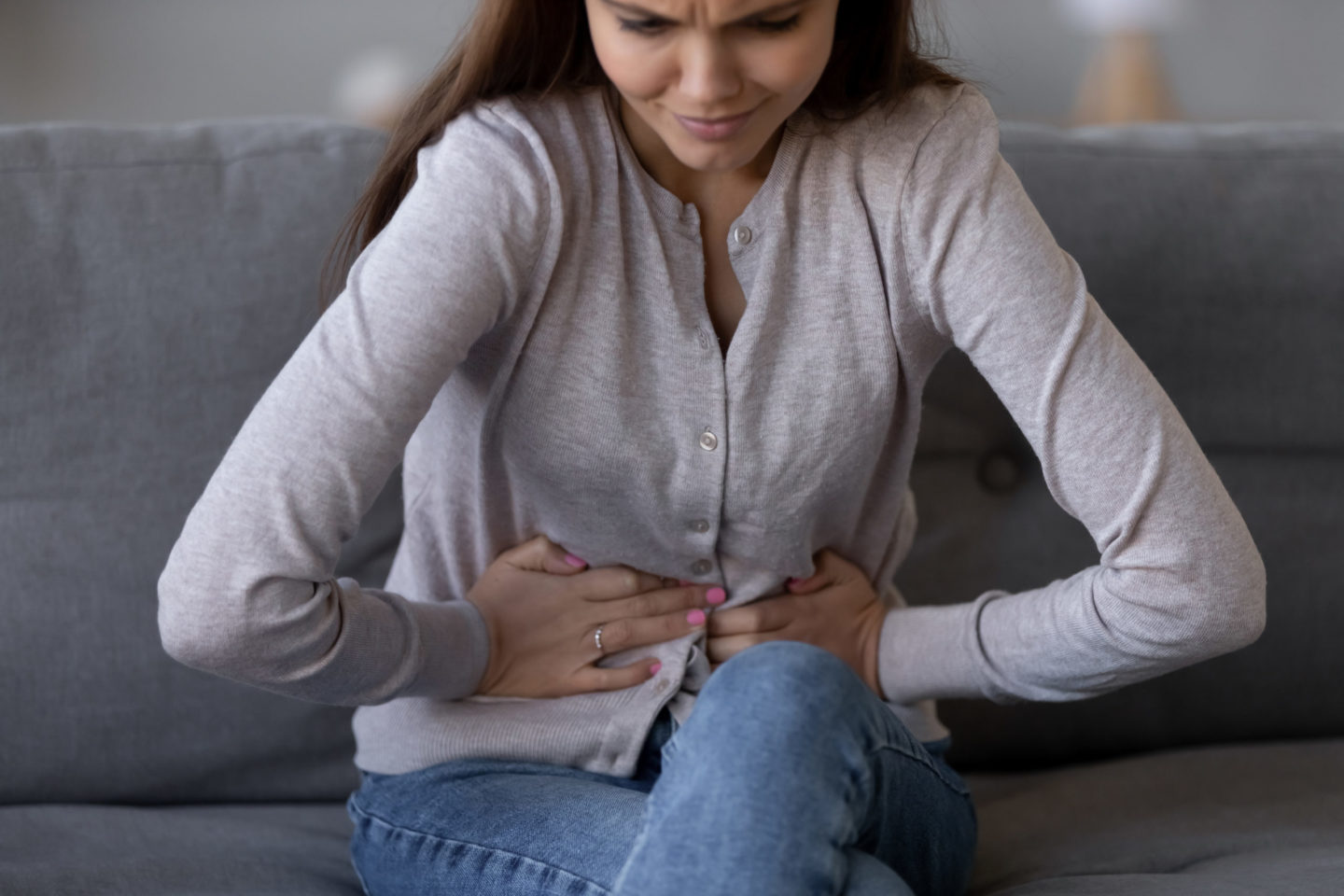 Eine Frau sitzt auf einem Sofa und hält sich den Bauch, sichtbar unter Schmerzen leidend. Das Bild veranschaulicht typische Symptome von Reizdarm, die mit Bauchschmerzen und Unwohlsein verbunden sind.