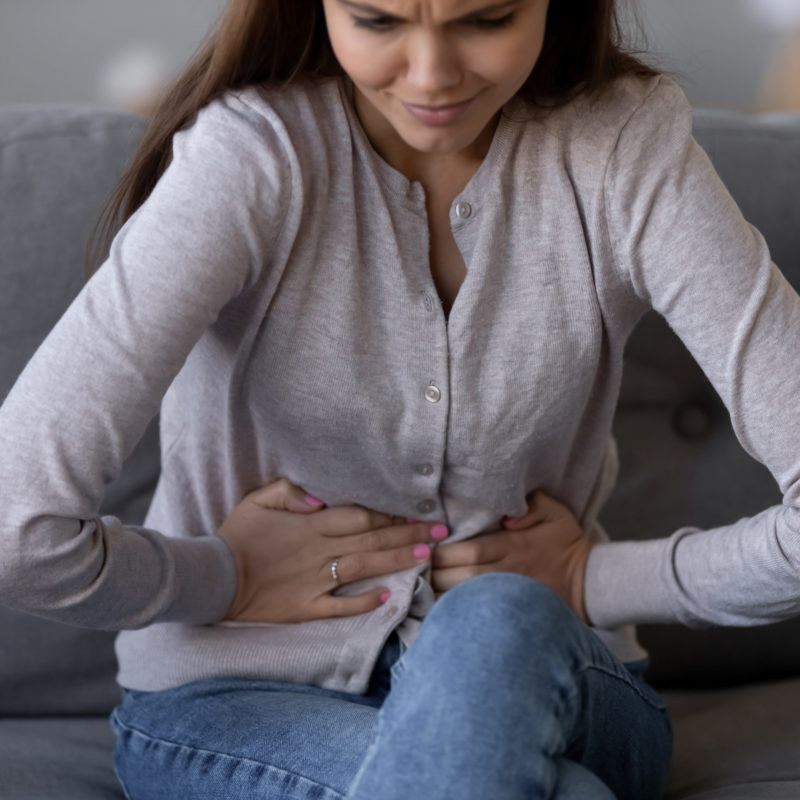 Eine Frau sitzt auf einem Sofa und hält sich den Bauch, sichtbar unter Schmerzen leidend. Das Bild veranschaulicht typische Symptome von Reizdarm, die mit Bauchschmerzen und Unwohlsein verbunden sind.