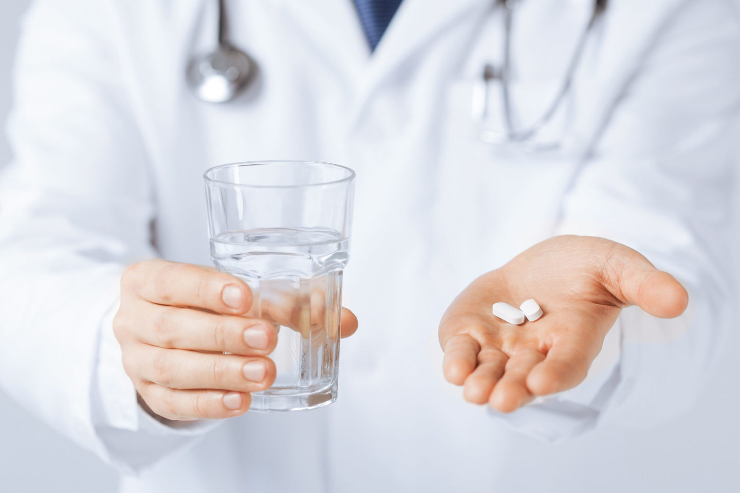 Ein Arzt in weißem Kittel hält ein Glas Wasser in einer Hand und zwei Tabletten in der anderen Hand. Die Abbildung symbolisiert die Medikation für Patienten mit Colitis ulcerosa und Morbus Crohn.