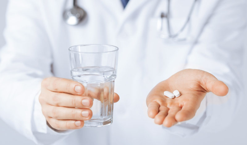 Ein Arzt in weißem Kittel hält ein Glas Wasser in einer Hand und zwei Tabletten in der anderen Hand. Die Abbildung symbolisiert die Medikation für Patienten mit Colitis ulcerosa und Morbus Crohn.