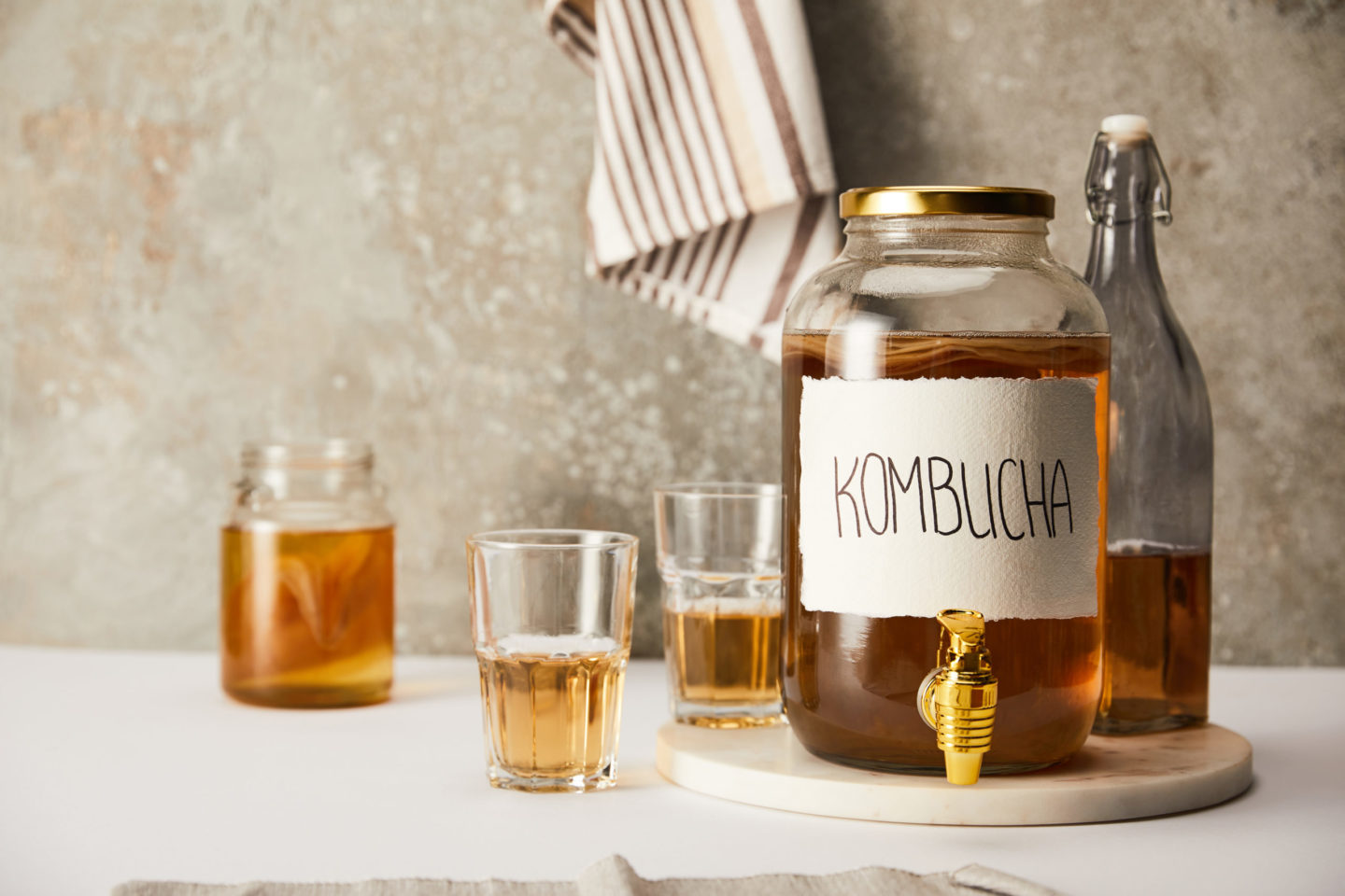 Ein Glasbehälter mit Kombucha, einem fermentierten Teegetränk, steht auf einem Tisch. Daneben befinden sich weitere Gläser mit Kombucha und ein Flasche im Hintergrund. Das Bild vermittelt eine gesunde Lebensweise und die Bedeutung der Darmflora.