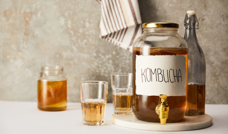 Ein Glasbehälter mit Kombucha, einem fermentierten Teegetränk, steht auf einem Tisch. Daneben befinden sich weitere Gläser mit Kombucha und ein Flasche im Hintergrund. Das Bild vermittelt eine gesunde Lebensweise und die Bedeutung der Darmflora.