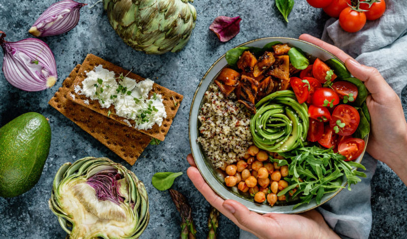 Schüssel mit buntem vegetarischem Salat, gefüllt mit Quinoa, Kichererbsen, Gurkenspiralen, Cherry-Tomaten, Rucola und geröstetem Gemüse, umgeben von frischen Zutaten wie Avocado, Artischocken und roten Zwiebeln. Ideal für eine fleischfreie Ernährung.