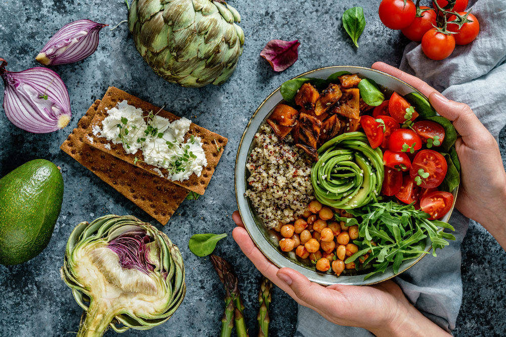 Schüssel mit buntem vegetarischem Salat, gefüllt mit Quinoa, Kichererbsen, Gurkenspiralen, Cherry-Tomaten, Rucola und geröstetem Gemüse, umgeben von frischen Zutaten wie Avocado, Artischocken und roten Zwiebeln. Ideal für eine fleischfreie Ernährung.