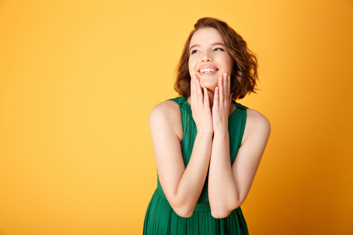 Junge Frau in grünem Kleid, die lächelt und ihre Wangen berührt, vor einem gelben Hintergrund, symbolisiert Freude und Wohlbefinden, passend zum Thema der Serotoninproduktion im Zusammenhang mit dem Darmmikrobiom.