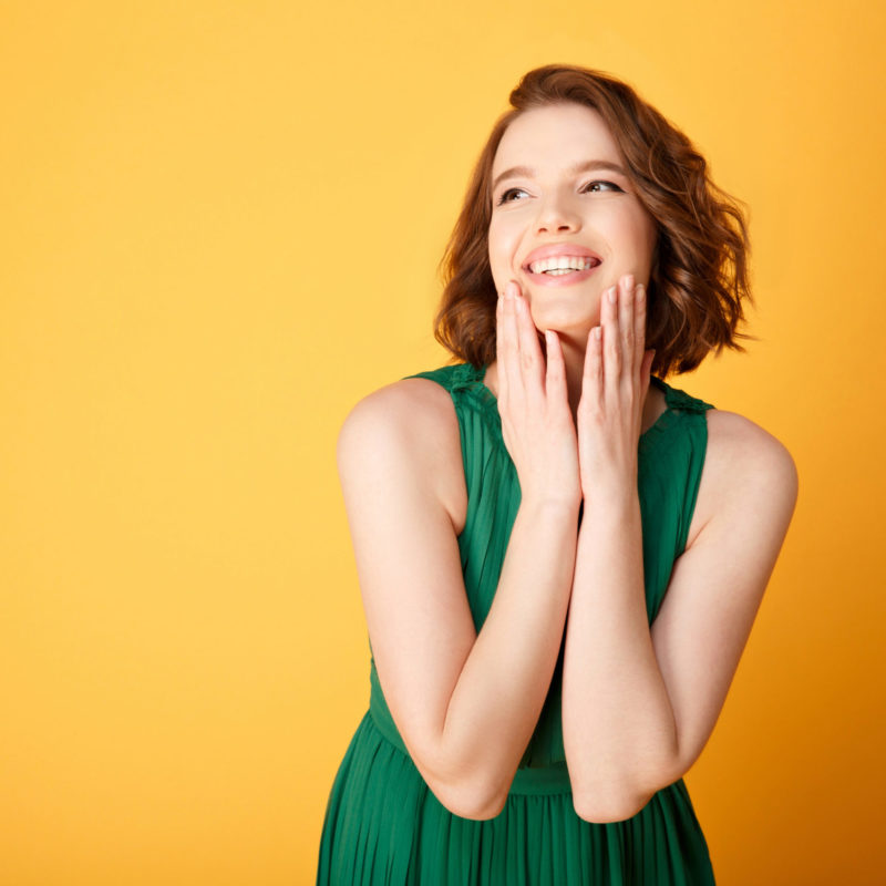 Junge Frau in grünem Kleid, die lächelt und ihre Wangen berührt, vor einem gelben Hintergrund, symbolisiert Freude und Wohlbefinden, passend zum Thema der Serotoninproduktion im Zusammenhang mit dem Darmmikrobiom.