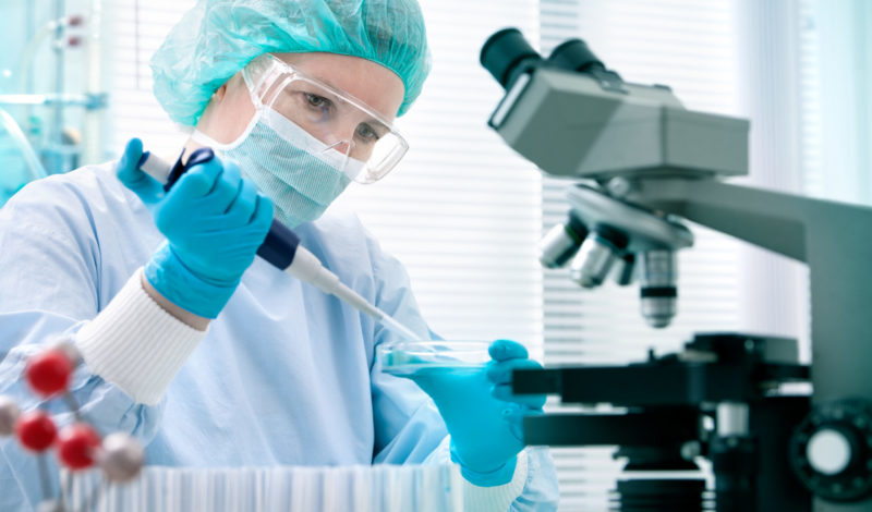 Wissenschaftlerin im Labor, die mit einer Pipette Proben in eine Petrischale überträgt, während ein Mikroskop im Hintergrund steht. Fokus auf DNA-Tests und Darmflora-Forschung.