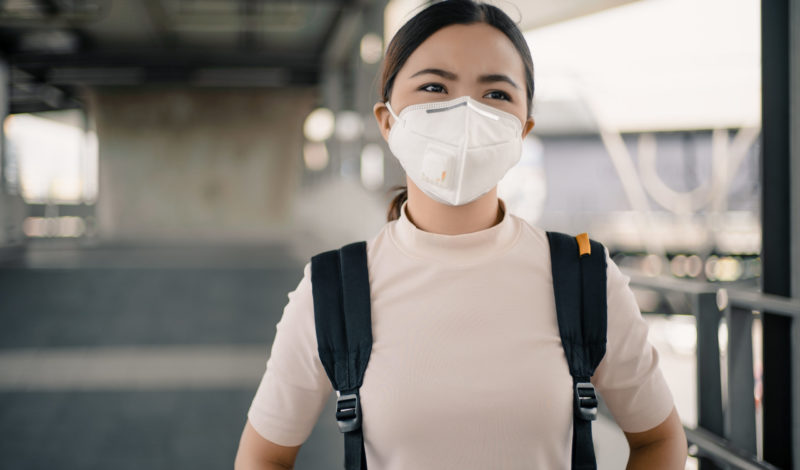 Frau mit Schutzmaske steht in einem modernen urbanen Umfeld, was auf die Bedeutung von Gesundheitsmaßnahmen während der COVID-19-Pandemie hinweist.