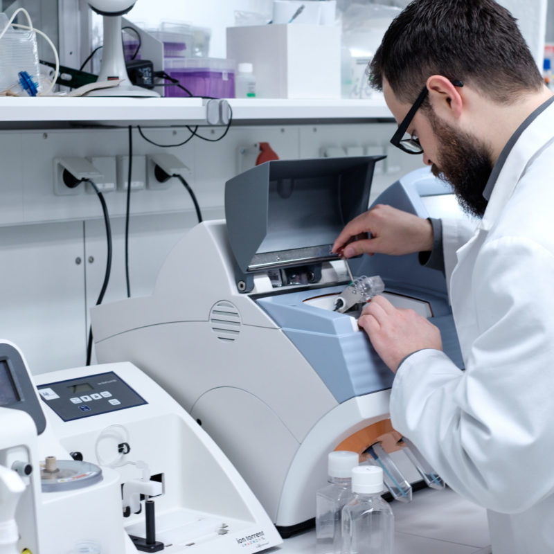 Wissenschaftler im Labor arbeitet an einem modernen Analysegerät, um Experimente durchzuführen. Im Hintergrund sind verschiedene Laborausrüstungen und Behälter sichtbar.