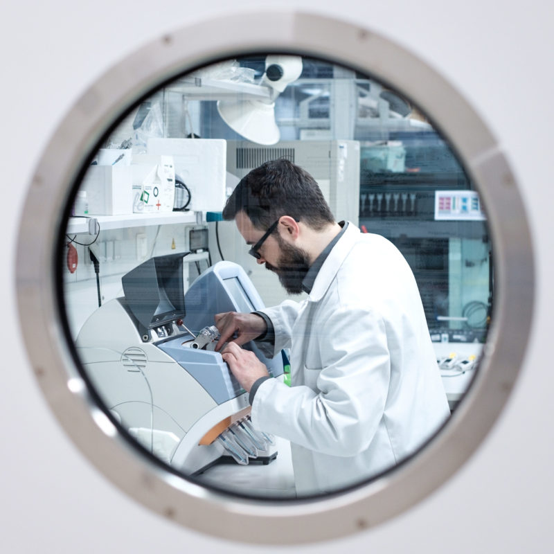 Laborant arbeitet an einem modernen Analysegerät in einem Labor, sichtbar durch ein rundes Fenster. Die Ausstattung im Hintergrund deutet auf eine hochtechnisierte Umgebung hin.