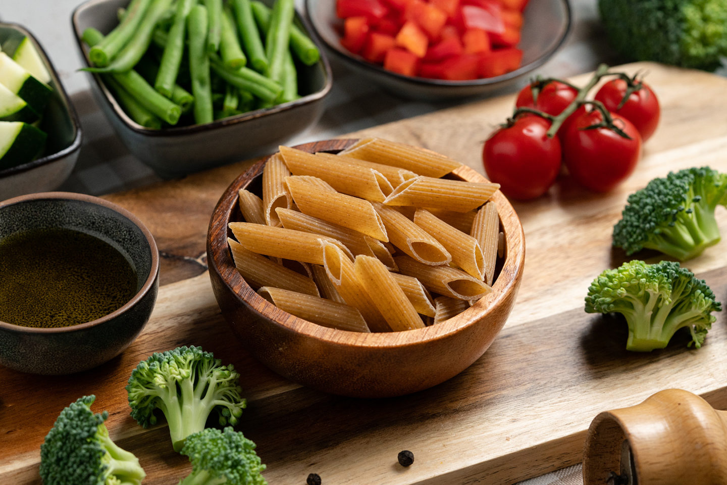 Vollkorn-Pasta in einer Holzschüssel umgeben von frischem Gemüse wie Brokkoli, Zucchini, grünen Bohnen, roten Paprika und Kirschtomaten sowie Olivenöl auf einem Holzbrett. Ideal für gesunde Rezepte zur Unterstützung der Darmbakterien.