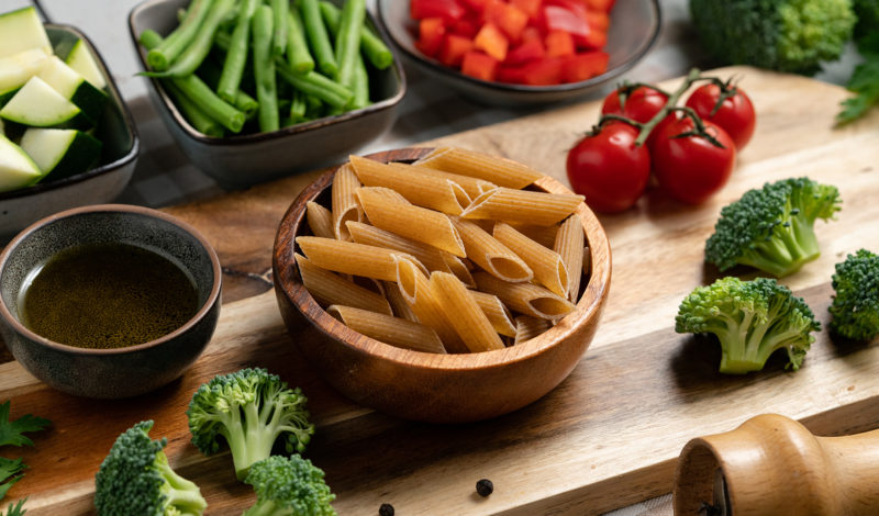 Vollkorn-Pasta in einer Holzschüssel umgeben von frischem Gemüse wie Brokkoli, Zucchini, grünen Bohnen, roten Paprika und Kirschtomaten sowie Olivenöl auf einem Holzbrett. Ideal für gesunde Rezepte zur Unterstützung der Darmbakterien.
