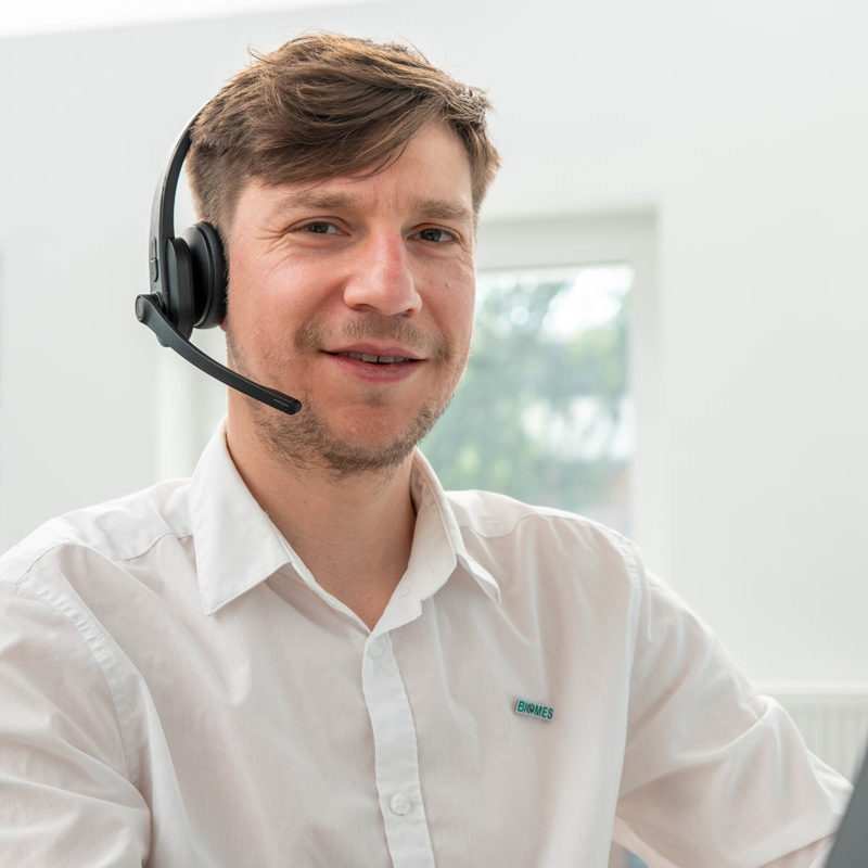 Mann mit Headset lächelt freundlich, sitzt an einem Schreibtisch in einem hellen Büro, ideal für Support- oder Beratungsdienste zu Darmbakterien.