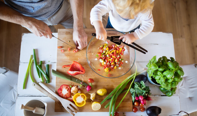 Ein Erwachsener und ein Kind bereiten gemeinsam einen bunten Salat aus frischem Gemüse auf einem Holzbrett zu. Verschiedene Gemüsesorten wie Paprika, Radieschen und Frühlingszwiebeln sind sichtbar, während das Kind mit einem Löffel in einer Schüssel rührt.