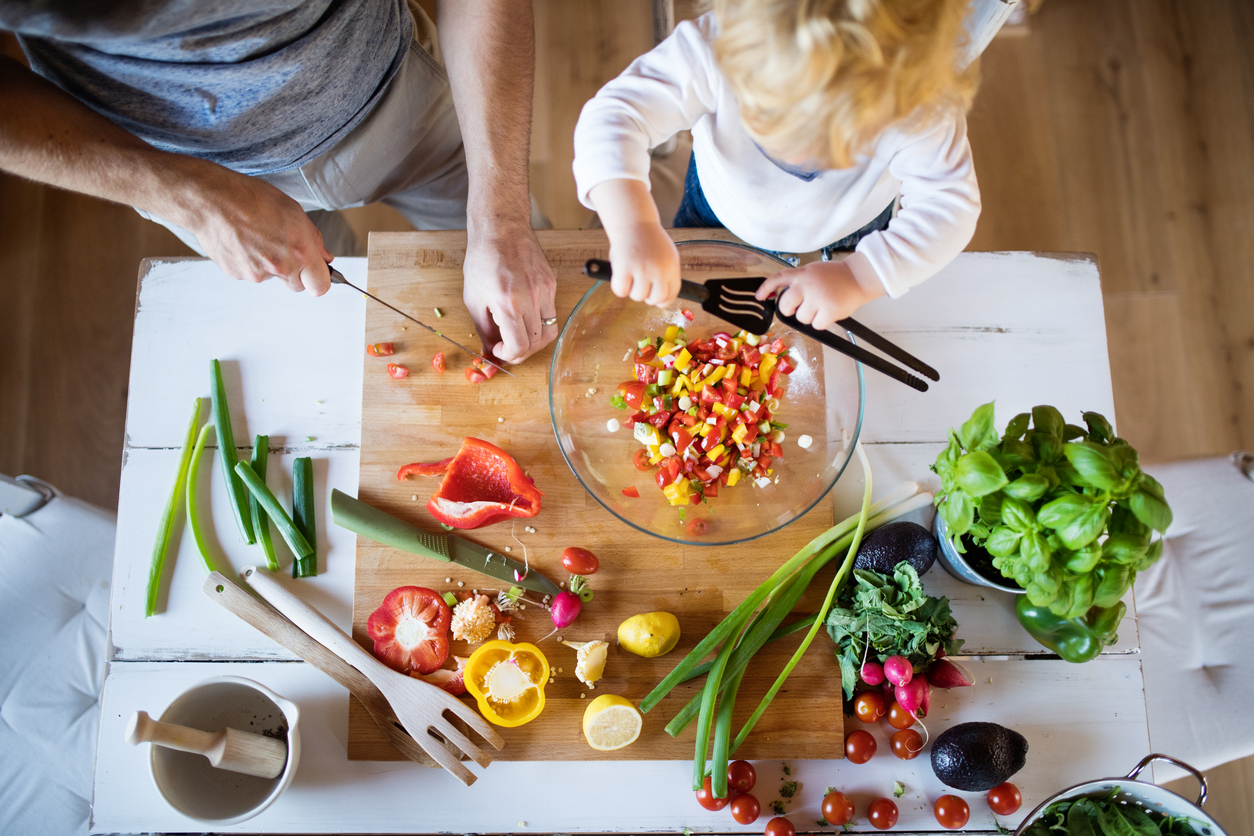 Ein Erwachsener und ein Kind bereiten gemeinsam einen bunten Salat aus frischem Gemüse auf einem Holzbrett zu. Verschiedene Gemüsesorten wie Paprika, Radieschen und Frühlingszwiebeln sind sichtbar, während das Kind mit einem Löffel in einer Schüssel rührt.