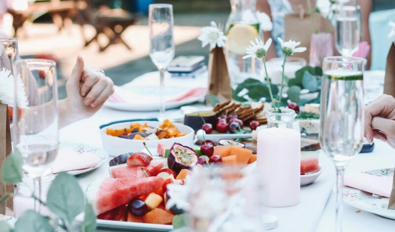 Ein festlich gedeckter Tisch mit frischem Obst, darunter Wassermelonen, Trauben und exotische Früchte, umgeben von Gläsern Sekt und dekorativen Elementen wie Kerzen und Blumen. Ideal für eine Sommerfeier oder ein Picknick.