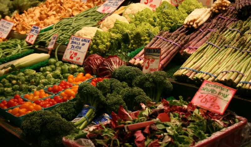 Bunte Auswahl an frischem Gemüse auf einem Marktstand, einschließlich Salat, Brokkoli, Tomaten und Kräutern, mit Preisschildern.