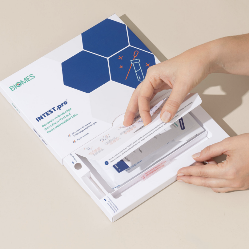 Hand, die ein Testkit aus einer Verpackung mit hexagonalem Design entnimmt, auf einem neutralen Hintergrund. Das Kit wird für einen medizinischen oder diagnostischen Test verwendet.