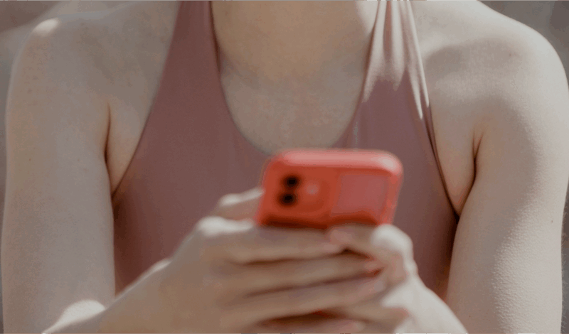 Junge Person in Sportkleidung hält ein rotes Smartphone in den Händen, während sie im Freien sitzt.